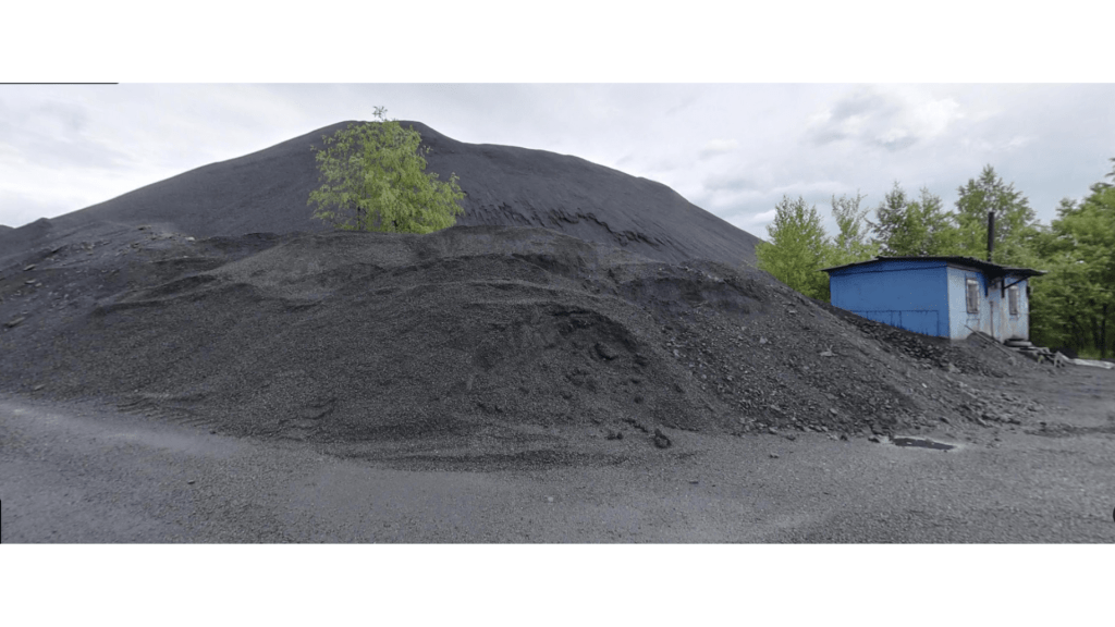 A view of a shungite mining site featuring a large mound of black material, with a small blue building and some greenery in the foreground.
