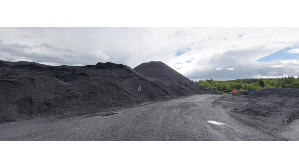 A panoramic view of a shungite mining site, featuring large black piles of shungite and a gravel road under a cloudy sky.