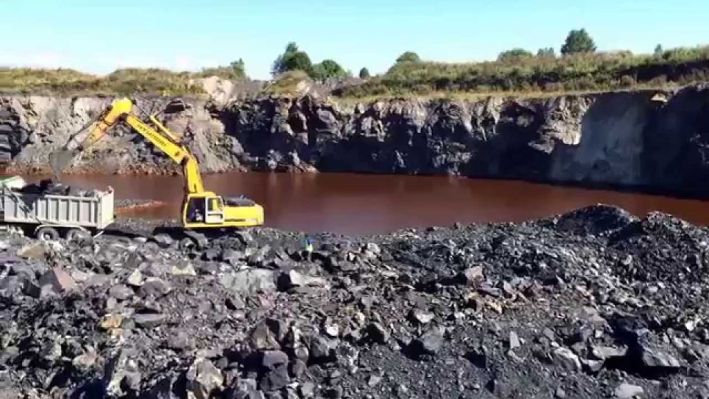 A yellow excavator loading rocks into a truck at a shungite mining site, with a body of brown water in the background and rocky terrain surrounding the area.